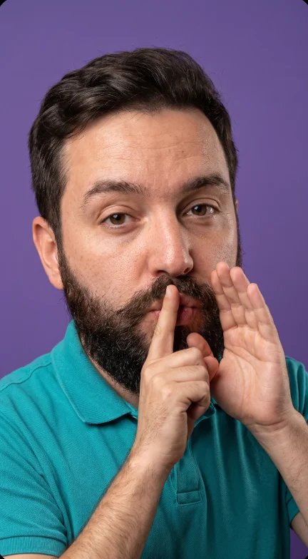 Selfie close-up at a home desk, evening lamp light. He leans in, finger to lips.